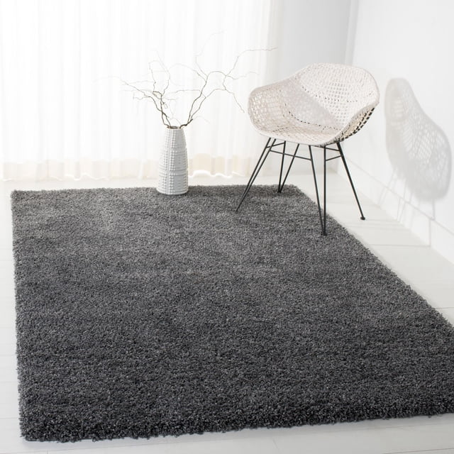 Safavieh California Solid Plush Shag Area Rug in dark grey with a super dense 4.5kg/m² polypropylene pile, placed on a white floor next to a white woven chair with black metal legs and a white ceramic vase holding bare branches, illuminated by natural light through sheer curtains, often used to add plush comfort and texture to modern living spaces.