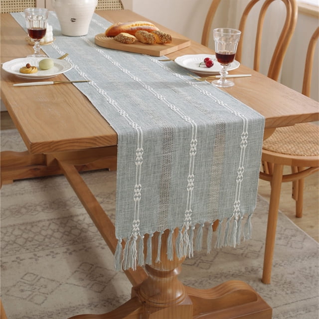 Dvkptbk Boho Table Runner in rustic linen with macrame tassels, shown in a light blue color with a woven texture and intricate lace patterns. The table runner measures 38x183cm (14.96x72in) and is displayed on a wooden dining table with a Bohemian-style setting, featuring bread, glassware, and plates. The design includes hollowed-out details and fringe lace, ideal for Bohemian home decor, dining, or bedroom table decoration.