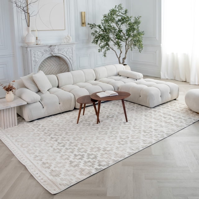 Rug Branch Astra Modern Indoor Area Rug in beige cream with abstract Moroccan pattern, on light wood flooring in a bright living room featuring a tufted cream sectional sofa, nested wooden coffee tables with an open book, a white marble fireplace, and a tall green plant near white paneled walls and sheer curtains.