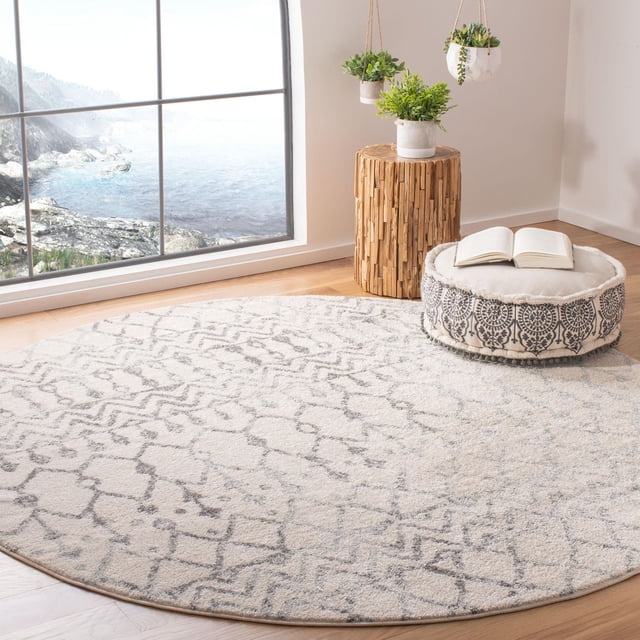 SAFAVIEH Tulum Glen Moroccan Geometric Area Rug in a bright room with coastal views through a large window. This round accent rug features soft cream and gray rustic geometric patterns on light hardwood floors. It is styled with a wooden stump side table holding a green potted plant, a patterned round floor cushion with an open book, and hanging planters. The rug’s soft synthetic yarns add comfort and boho-chic character, also suitable for creating a cozy reading nook.