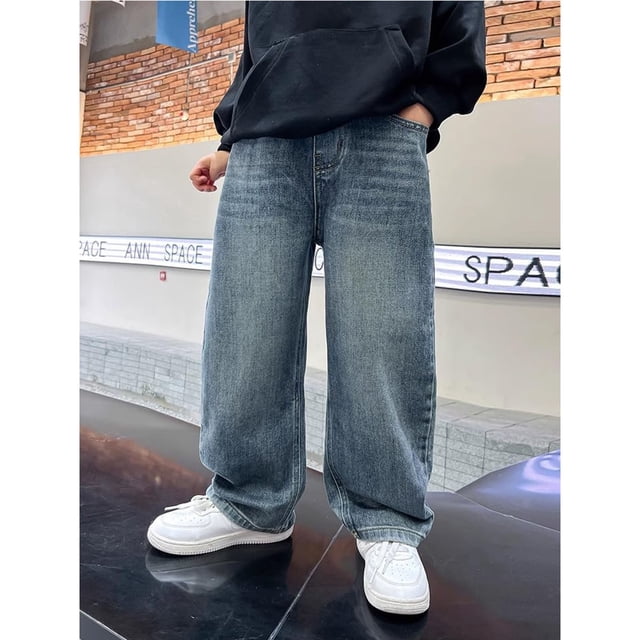 PMVFHDE Jeans Pantalon Garçon pour Garçons avec Taille Élastique en denim Bleu, Montré en Coupe Ample. Ces Pantalons de Printemps/automne pour Enfants Sont Faits de Tissu polyester Doux et Respirant. les Jeans Présentent un style cargo Décontracté, Idéal pour le Quotidien, les Réunions de Famille Ou la Photographie de Rue. Exposés sur un Modèle Portant des Espadrilles Blanches et un Chandail à Capuchon Noir dans un Cadre Urbain.