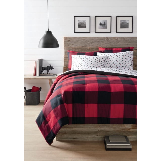 Canadiana Reversible 3pc Comforter Set on a rustic wooden bed featuring a red and black buffalo plaid comforter reversible to solid black. Includes two pillow shams with matching plaid and white reverse with small black and red tree prints. The room has white shiplap walls, three black-framed nature prints above the headboard, a black pendant light, a wooden side table with books and a black moose figurine, and a grey felt basket. Made of 100% polyester, machine washable, designed for warmth and year-round comfort.