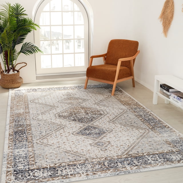 Rug Branch Atlas Persian Indoor Area Rug in beige grey with distressed abstract geometric patterns, on light wood flooring in a bright room with a rust-coloured wooden armchair, woven basket plant, and white side table with books, suitable for living or dining areas.