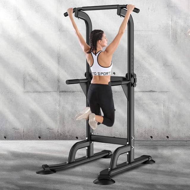 SOGES Height Adjustable Power Tower Dip Stands Pull up Bar Strength Training for Home Gym in black. The equipment features a heavy-duty metal frame with a splayed anti-eversion base for stability. It includes adjustable height settings from 65 to 82.7 inches and a weight capacity of 330 lbs. The image shows a person performing a pull-up in a home gym setting with a concrete wall background.