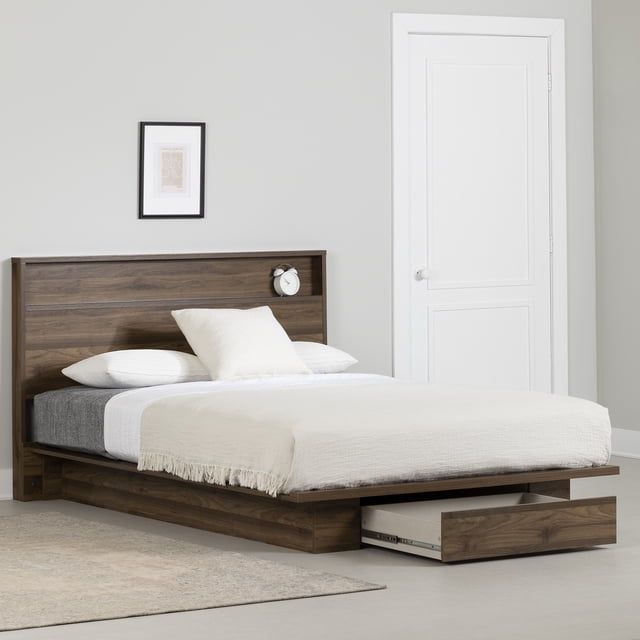 South Shore Musano Full/Queen Platform Bed and Headboard Set in natural walnut finish with a low-profile platform frame featuring a full-extension storage drawer at the foot and a headboard shelf holding a white alarm clock. The bed is dressed in white and beige bedding, set in a minimalist room with a light grey wall, framed abstract art, and a white door. This urban modern bed supports up to 500 lb and does not require a box spring, suitable for maximizing bedroom storage.