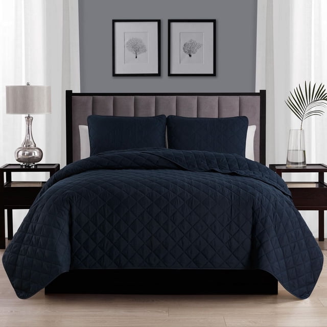 Swift Home Diamond Quilt Set in dark navy blue on a bed with a gray upholstered headboard, featuring diamond quilted polyester fabric and matching pillow shams with 2-inch hems. The set adds comfort and style to a modern bedroom with dark wood nightstands, a silver lamp, and framed botanical prints.