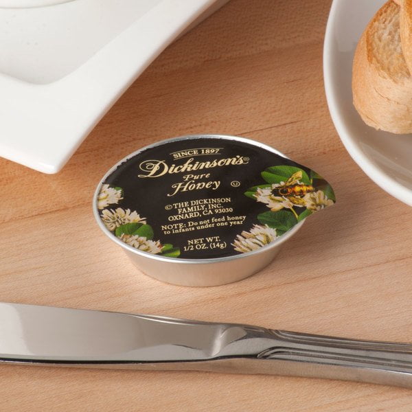 Dickinson's Pure Honey .5oz portion cup displayed on a wooden table. The cup features elegant black and gold packaging with floral graphics, emphasizing its premium clover honey content. Positioned near a bread roll, knife, and white plate, ideal for breakfast or in-room dining.