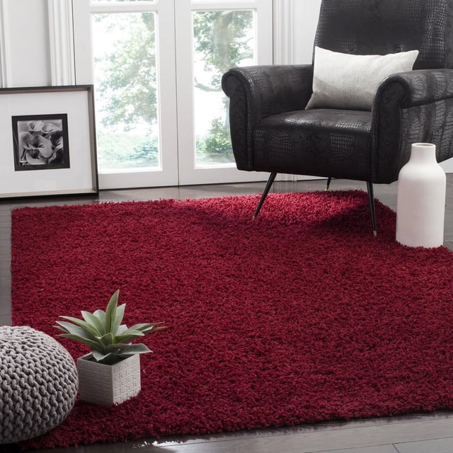 Safavieh Athens Solid Plush Shag Area Rug in deep red on dark hardwood floor, featuring a thick polypropylene plush pile. Styled with a black textured armchair with white lumbar pillow, white ceramic vase, gray knitted pouf, and a small green succulent in a white square pot by a bright window with white trim. Often used to add warmth and softness to living rooms.