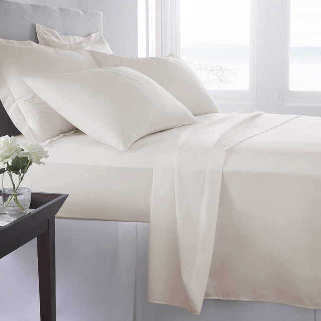 Johnson Home Ensembles de Draps en Coton Peigné 500 Fils de Couleur Crème sur un Lit Fait, Comprenant un Tissu en Coton Finement Tissé, Respirant et durable avec Drap-Housse, Drap Plat et Taies d'Oreiller, Montrés dans une Chambre Éclatante avec une Fenêtre et une table d'Appoint Tenant des Fleurs Blanches.