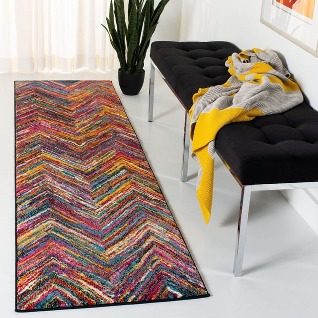 SAFAVIEH Aruba Tapis à Chevrons Bernadine avec un Motif à Chevrons Multicolore Vif en Rose, Jaune, Bleu et orange, Tissé Mécaniquement à Partir de Fils Synthétiques Durables, Posé sur un Plancher Blanc à Côté d'Un Banc Capitonné Noir avec des Pattes Chromées et un Jeté Jaune-Gris, Près d'Une Grande Plante Serpent Verte et de Voilages Blancs, Idéal pour Ajouter de la Couleur aux Espaces de Vie Modernes.