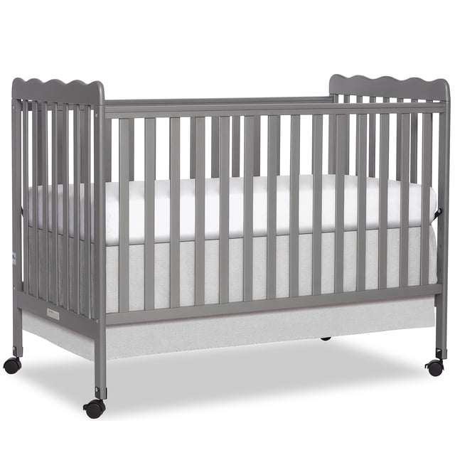 Dream On Me Carson Classic 3-in-1 Convertible Crib, Model #675, in grey with vertical slats and scalloped top rails. It includes a white mattress and light grey fabric skirt, with four black safety locking wheels for easy mobility. Made from sustainable New Zealand pinewood, it features a stationary rail design and measures 54L x 31W x 40H inches, ideal for nursery furniture.