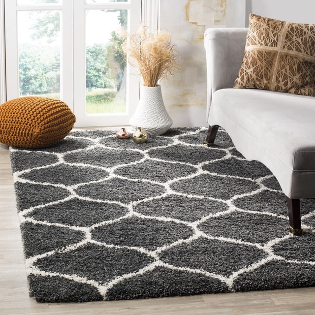Safavieh Hudson Tapis Shag Géométrique Arline avec une Épaisse Peluche Shag Gris Foncé et un Motif de Carreaux en Forme d'Oignon Blanc, Exposé sur un Plancher de Bois Clair à Côté d'Un sofa Gris Pâle avec des Coussins à Motif Brun, un vase Texturé Blanc avec des Herbes Séchées, et un pouf Tricoté orange Brûlé Près d'Une Porte en Verre, Convient pour Ajouter de la texture aux Espaces de Vie Modernes.