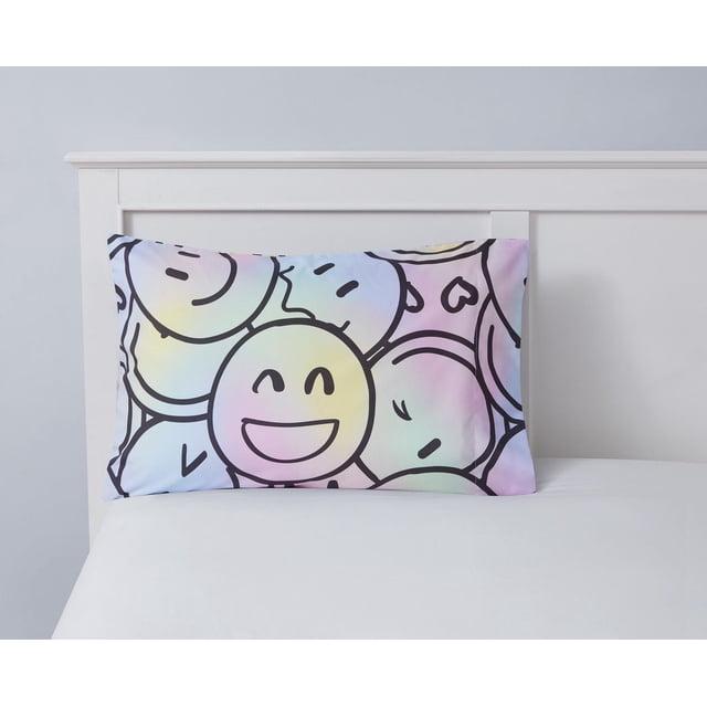Mainstays Kids Printed Pillowcase featuring black-outlined smiley faces with various expressions on a pastel gradient background in yellow, blue, pink, and green, made of 100% polyester microfiber, measuring 20 by 30 inches, displayed on a white bed with a white wooden headboard, suitable for adding playful decor to a child's bedroom and often used as a fun decorative accent.