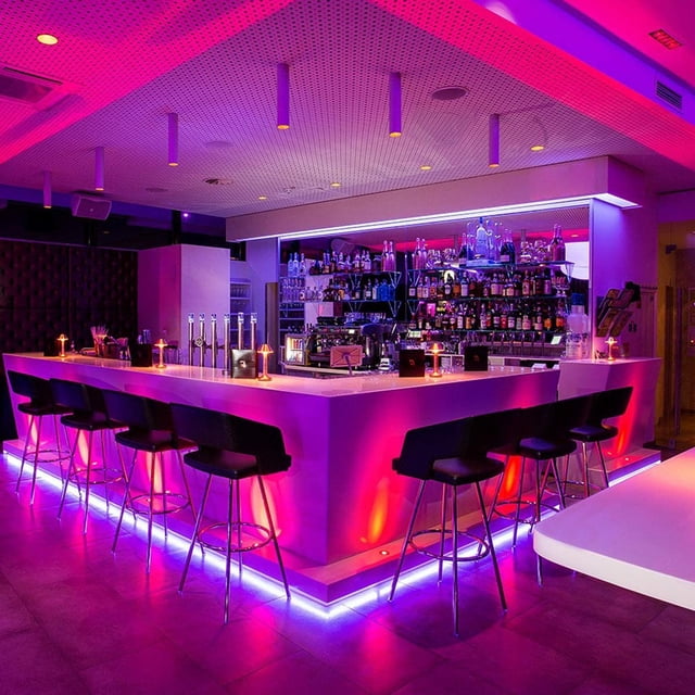 Strip Lights, 16.4ft RGB LED Light Strips Kit with Remote Control, showcased in a modern bar environment. The LED strips provide vibrant, color-changing illumination in pink, purple, and blue tones, enhancing the bar counter and seating area. Ideal for novelty lighting, bedroom, kitchen, TV, or event decoration.