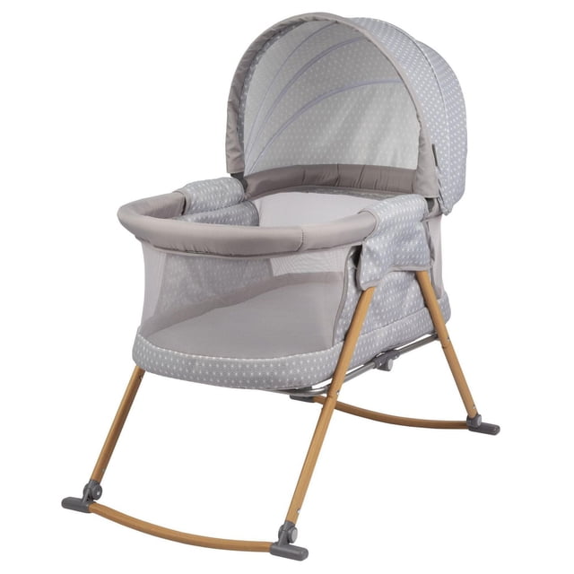 Safety 1st Amherst Bassinet in light grey with white star pattern, featuring breathable mesh sides, padded rim, and retractable canopy. It has wooden rocking legs with grey feet, ideal as a bedside bassinet for infant sleep up to 20 lbs.
