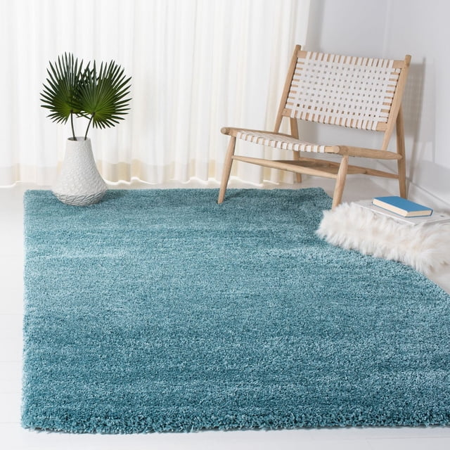 Safavieh Milan Harlow Solid Shag Area Rug in radiant ivory with a plush, thick texture, shown in a minimalist room with white walls and sheer curtains. The rug is under a wooden lounge chair with white woven straps, beside a white vase with green palm leaves and a white faux fur cushion with books, adding warmth and contemporary style.