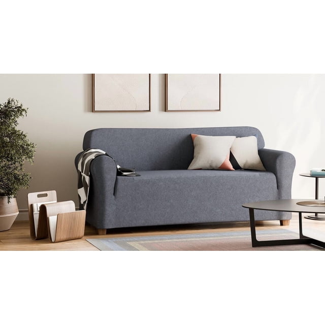 SmartFit Chenille Stretch Sofa Recycled Fabric Slipcover in dark grey fitted on a three-seat sofa with rounded arms and wooden legs, made with soft, durable chenille fabric containing 20% recycled polyester. The one-piece slipcover protects the sofa from dirt, pet hair, and stains, shown with two decorative pillows, a black and white throw, wooden magazine holder, potted plant, and minimalist wall art in a modern living room.