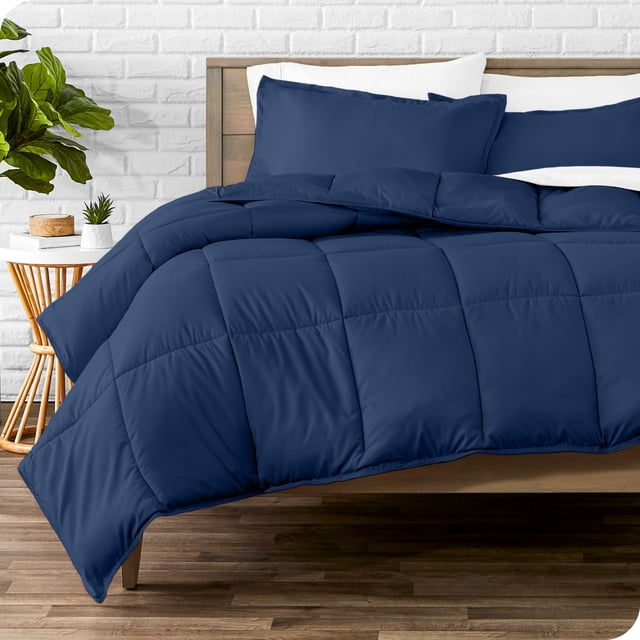 Bare Home Goose Down Alternative Comforter Set - 2 Piece Set - Twin/Twin XL, Dark Blue. The comforter set includes a dark blue quilted comforter (66” by 92”) and a matching pillow sham (20” by 26”). Made from hypoallergenic, ultra-soft brushed microfiber polyester, it provides breathable, all-season comfort. The box-stitch design ensures the fill stays in place. Shown on a wooden bed frame in a modern bedroom with a white brick wall and a side table with decor.