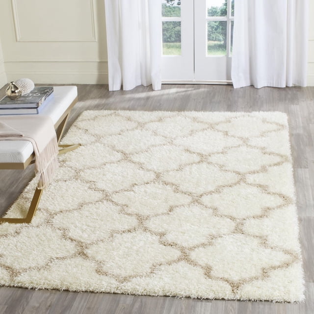 Safavieh Montreal Lennart Geometric Shag Area Rug in cream with beige quatrefoil pattern, featuring a plush shag pile texture on a light wood floor in a bright room with white curtains and a bench with books and a throw, ideal for contemporary or transitional spaces.