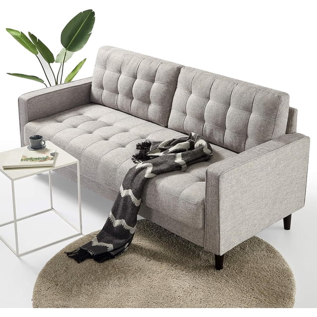 Zinus Benton 3-Seat Upholstered Grid Tufted Sofa Couch, Soft Grey in a front angled view with soft grey polyester upholstery and grid tufting on seat and back cushions, supported by dark wooden legs, placed on a round beige rug with a black and white throw blanket on the seat, beside a white metal side table with a cup and books, and a green plant behind.