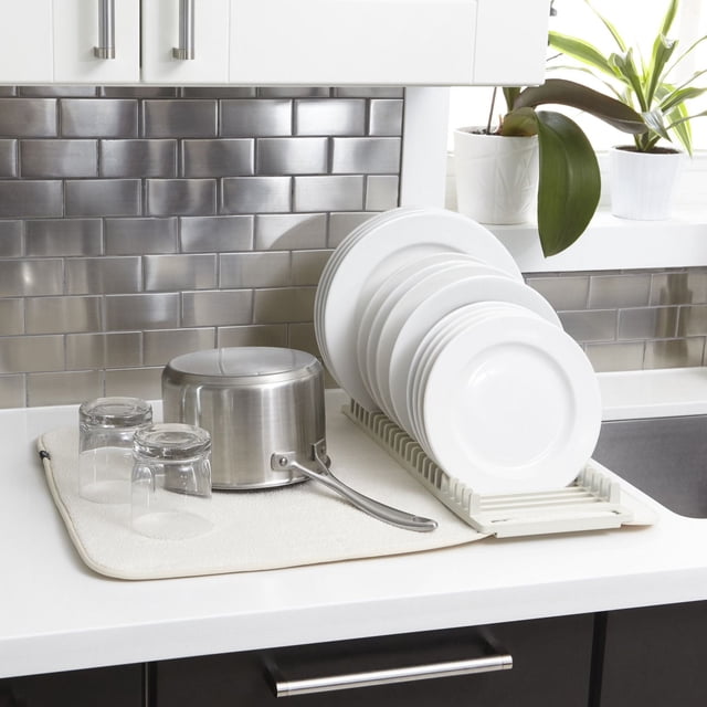 Umbra UDry Tapis d'Égouttage pour Vaisselle en Lin sur un Comptoir de Cuisine Blanc avec un pot en Acier Inoxydable, Deux Verres Transparents Inversés et un Égouttoir Blanc Tenant des Assiettes Blanches. le Tapis en Microfibre Absorbe l'Eau et Protège le Comptoir des Égratignures et de l'Humidité, contre un Dosseret de Carreaux de Métro en Acier Inoxydable avec des Armoires Blanches et des Plantes en pot Vertes à Proximité. le Tapis Se Plie de Façon Compacte pour un Rangement Facile.