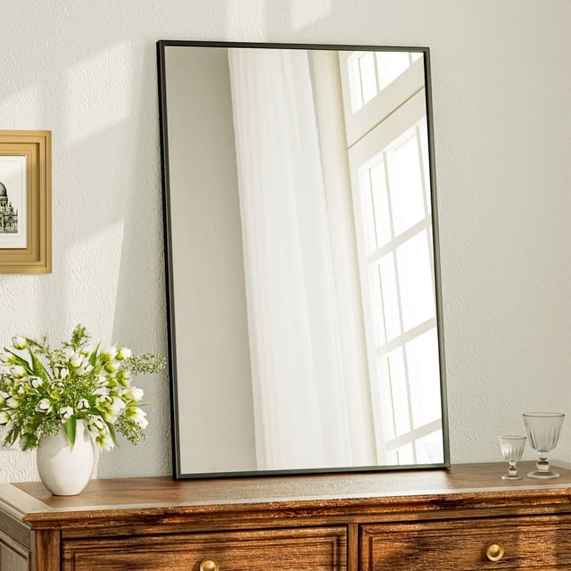 BEAUTYPEAK 24"x36" bathroom mirror with a rectangular black metal frame, displayed leaning against a wall on a wooden dresser. The mirror features a slim aluminum alloy frame and high-definition shatter-proof glass, reflecting a bright window and curtain. Suitable for bathrooms, bedrooms, living rooms, and hallways, it offers versatile mounting options for vertical or horizontal installation.
