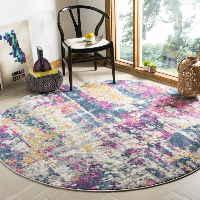 SAFAVIEH Madison Ivory Abstract Area Rug, round with a multicolored abstract pattern in ivory, pink, yellow, and blue tones, placed on a light wood floor near a large window. A black wooden chair with a white cushion sits on the rug, alongside a gray knitted pouf holding books and a decorative item, with a woven lantern and small potted plants by the window, suitable for enhancing modern and transitional home interiors.