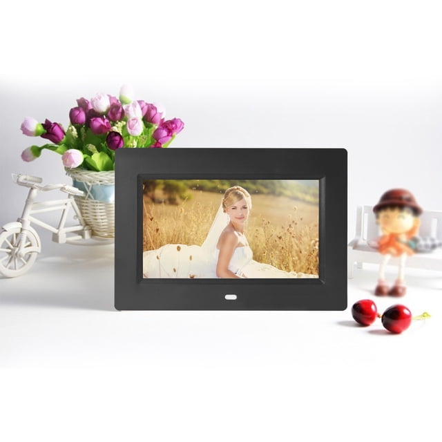 Dvkptbk Ultra-Thin Fashion High-Definition LED Electronic Photo Frame Multimedia Advertising Player 7 Inch Digital Photo Frame in black. The frame features a 7-inch LED LCD screen with 800x480 resolution, supporting up to 1200P pictures. It includes functions like picture browsing, MP3/MP4 playback, background music, clock, alarm clock, and a perpetual calendar. The frame is displayed on a white surface with decorative elements like a basket of flowers, a miniature bicycle, and figurines in the background.