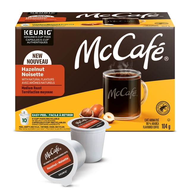 McCafé Premium Hazelnut Flavoured, Medium Roast, K-Cup Coffee Pods box with 10 recyclable pods compatible with Keurig machines, displayed alongside a clear glass mug of steaming medium roast coffee and whole hazelnuts, highlighting natural flavours and 100% Rainforest Alliance Certified Arabica beans.