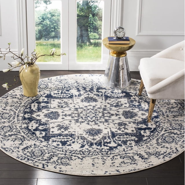 Safavieh Madison Katina Traditional Faded Area Rug, round with a blue and white faded medallion pattern, power loomed for durability, placed on dark hardwood flooring in a bright room with white walls and glass doors. The rug suits transitional decor and is shown with a beige upholstered chair, a metallic side table with a gold top holding a clock and books, and a beige vase with white flowers. It is also suitable as a decorative accent rug in living spaces.