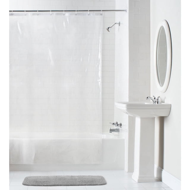 Mainstays Waterproof Heavyweight PEVA Shower Curtain Liner in clear translucent PEVA with rustproof metal grommets and reinforced mesh header, hanging inside a white tiled bathtub and shower area with a silver rod, beside a white pedestal sink and oval mirror, with a gray bath mat on white tile floor.