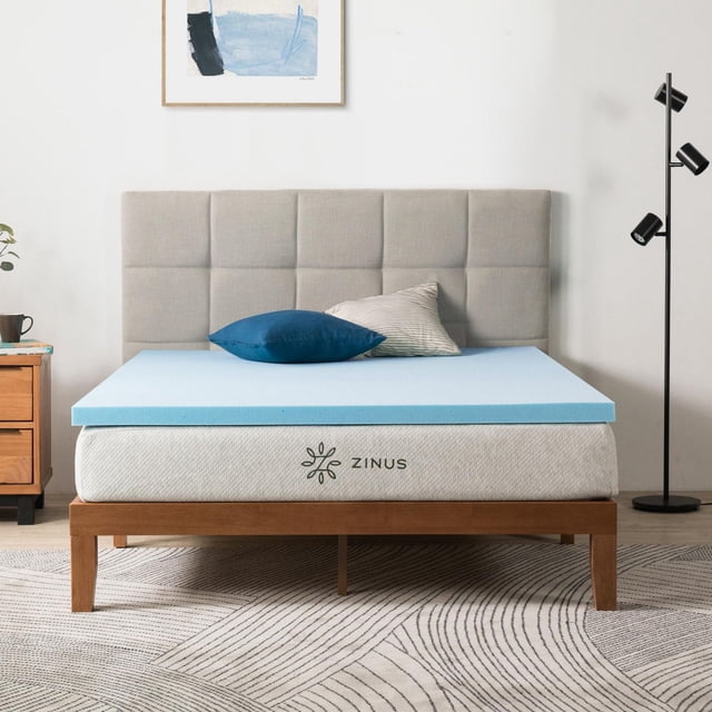 Zinus Spa Sensations 2 inch Cooling Gel Memory Foam Mattress Topper on a queen mattress with a light grey upholstered headboard. The blue gel foam topper lies flat on the white mattress cover branded with the Zinus logo. The wooden bed frame has a natural finish and stands on a patterned rug. Two pillows, one solid blue and one beige striped, rest on the topper. The topper is suitable for enhancing mattress comfort and cooling support.