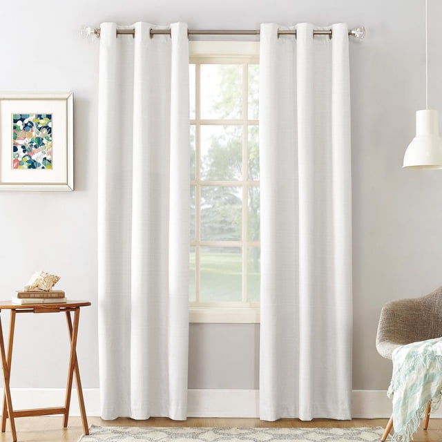 Mainstays Cooper Thermal Blackout Grommet Window Curtains Panel in white with subtle textured print, hanging on a silver rod with metal grommets. These energy-efficient curtains provide thermal insulation, noise reduction, and light blocking in a bright room with grey walls, wooden side table, framed abstract art, and beige armchair with a light blue throw.
