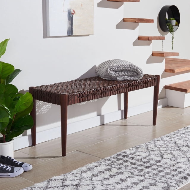 Safavieh Bandelier Cowhide Leather Bench with woven dark brown leather seat and tapered wooden legs, set in a modern entryway with a folded gray knit blanket on top, a green potted plant nearby, floating wooden stairs, and a gray and white patterned rug, ideal for extra seating or surface space.