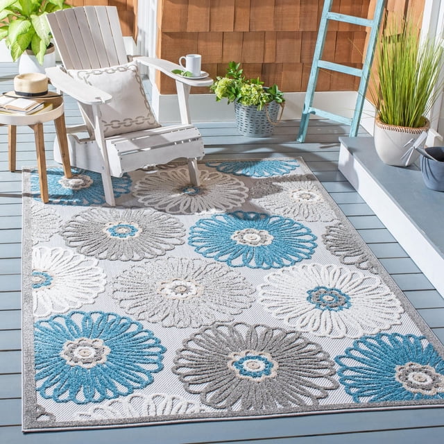 SAFAVIEH Cabana Anuki Floral Outdoor Area Rug on a grey wooden deck featuring large blue, grey, and white floral patterns. The durable outdoor rug is styled with a white Adirondack chair with a decorative pillow, a wooden side table with books, and potted plants, set against brown shingle siding and a blue ladder, perfect for patios or porches.
