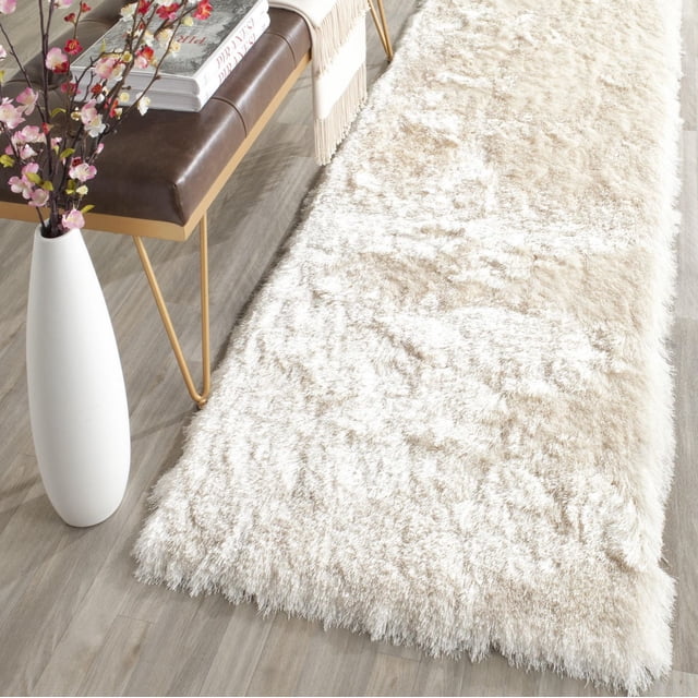 Safavieh Paris Darwin Plush Shag Area Rug in cream with thick, soft synthetic pile on light wood flooring beside a brown leather bench with gold legs and a white vase holding pink flowers, suitable for adding texture and warmth to modern home decor.