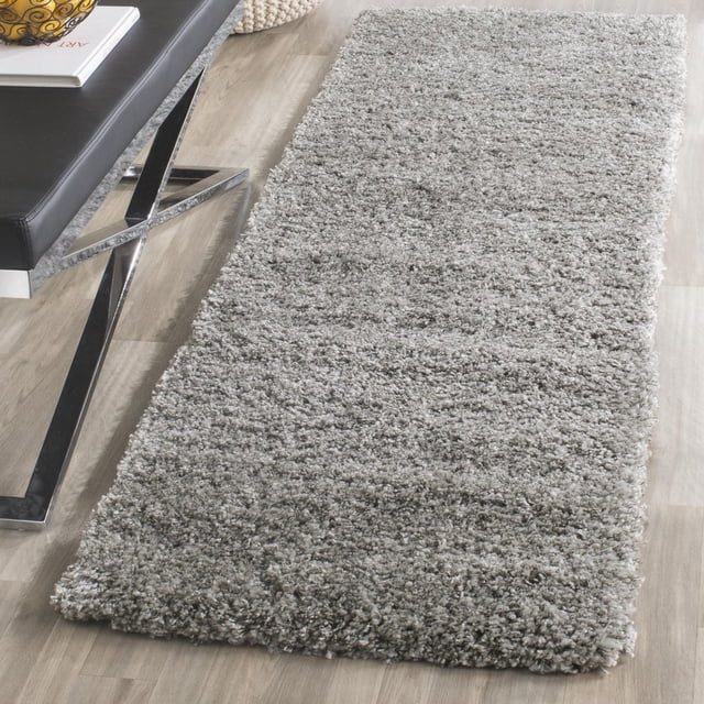 Safavieh California Solid Plush Shag Area Rug in grey with a dense 4.5kg/m² polypropylene pile, placed on a light wood floor next to a black leather bench with chrome legs, showcasing its plush texture and casual West Coast style, suitable for accent areas and layering over hard floors.