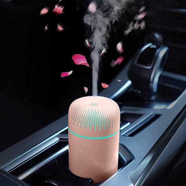 Car Diffuser Humidifier USB Aromatherapy Essential Oil Air Freshener in black, placed in a car cup holder. Emits cool mist with pink flower petals surrounding the mist. Compact portable humidifier with textured top, glowing LED light, and USB-powered functionality.