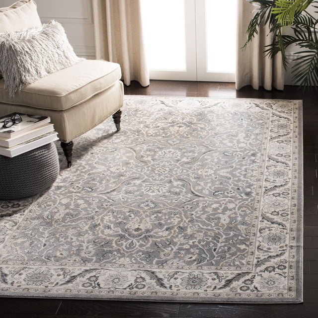 SAFAVIEH Isabella Kuzman Floral Bordered Area Rug featuring traditional floral patterns in soft greys and blues with a detailed border, power-loomed synthetic yarns in medium cut pile, displayed on dark hardwood flooring beside a beige armchair and grey knitted pouf, suitable for classic or casual room decor.