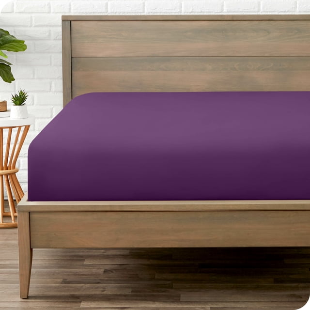 Bare Home Fitted Sheet from the Premium 1800 Collection in plum color, displayed on a twin bed. The ultra-soft microfiber sheet features deep pockets for mattresses up to 15 inches, is wrinkle-resistant, hypoallergenic, and suitable for luxury bedding. The bed is set in a minimalist room with a wooden frame and white brick wall.