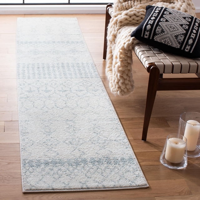 Safavieh Tulum Coriander Moroccan Geometric Area Rug runner in light blue and cream with a plush, hand-knotted wool texture, placed on a light hardwood floor next to a woven leather strap chair draped with a chunky cream knit throw and a black and white Moroccan-patterned pillow, accompanied by two white candles in glass holders on the floor.