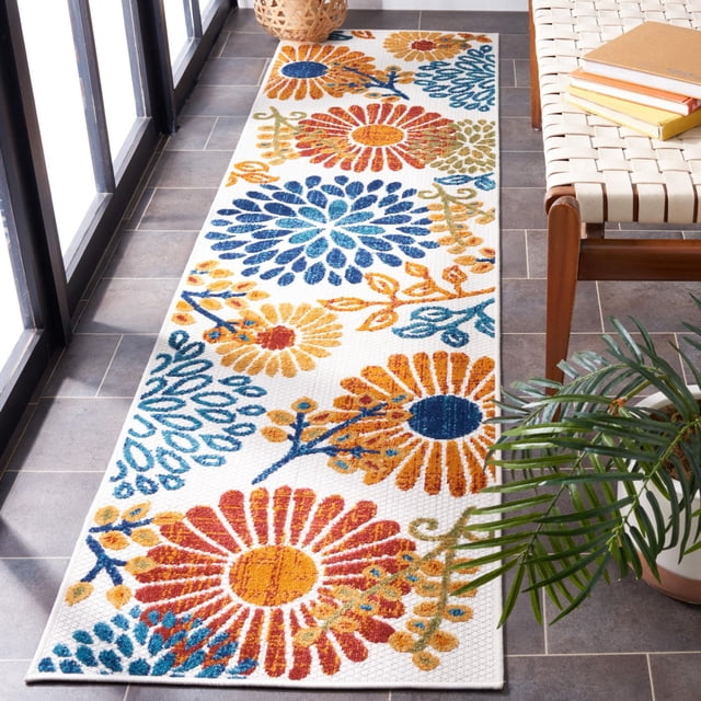 SAFAVIEH Cabana Garnet Floral Outdoor Area Rug with a rectangular shape and white background featuring large garnet red, orange, blue, and green floral and vine motifs. Placed on a tiled patio floor beside a woven bench with books and a potted plant, this durable outdoor rug is made from synthetic yarns resistant to weather, mold, mildew, and sun fading, suitable for decks, porches, and patios.