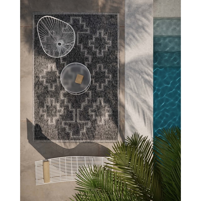ECARPET Esina Transitional Area Rug for living room, bedroom, dining room, entrance, hallway and more, with a dark neutral woven geometric pattern, displayed outdoors on a concrete patio next to a tiled pool, accompanied by white modern outdoor furniture and green palm leaves, often used to add texture and dimension to indoor or outdoor spaces.