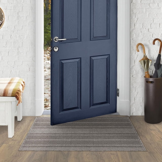 2 ft x 3 ft Olin Grey Mat placed inside a doorway on a wood floor with a navy blue paneled door partially open. This indoor doormat is machine tufted with durable polypropylene yarn in grey, featuring subtle horizontal stripes to protect floors from dirt, salt, and debris. The setting includes a white brick wall, a white bench with a plaid throw, and an umbrella stand with umbrellas. The mat is also suitable for entryways to reduce moisture damage.