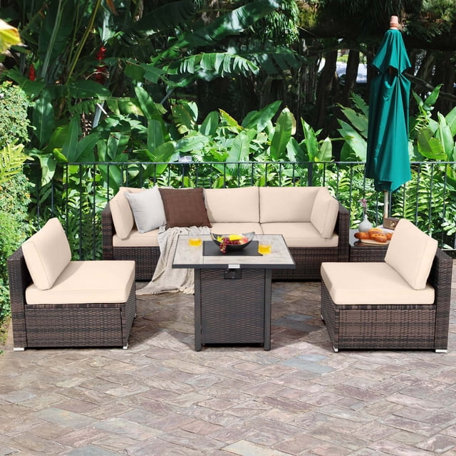 Costway 7PCS Patio Rattan Furniture Set featuring a 30-inch fire pit table with a ceramic tabletop, beige cushioned sofa seating, and a mix brown PE rattan frame. The set includes a fire pit table, coffee table, two corner sofas, and three armless sofas arranged on a stone patio with lush greenery in the background. The fire pit table provides 50,000 BTU heat output and comes with a waterproof PVC cover for durability.