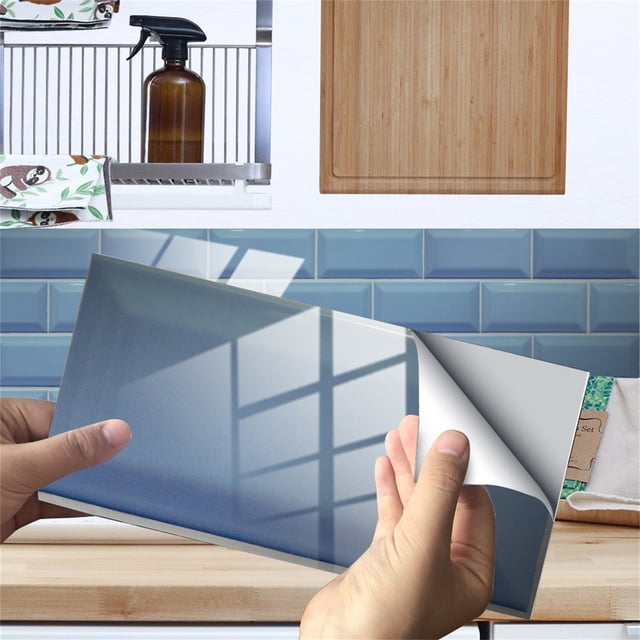 Vaidha Imitation Wall Tiles Crystal Film Tile Sticker Adhesive Floor Sticker with a glossy blue finish, resembling ceramic tiles, shown in a kitchen setting. A hand is peeling back the corner to reveal its adhesive PVC backing. Ideal for kitchen or bathroom walls, these decorative wall decals are easy to apply and suitable for flat surfaces like doors. Background includes a countertop and shelving with kitchen items.