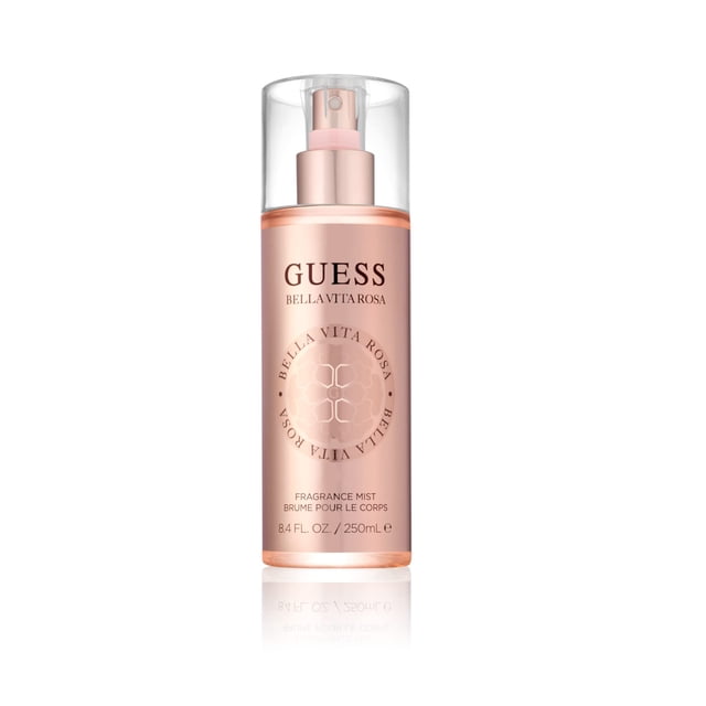 Guess Bella Vita Rosa Fragrance Mist, 8.4 Fl Oz, featuring a sleek cylindrical bottle with a soft pink hue and a transparent cap. The bottle displays the Guess logo and Bella Vita Rosa branding in elegant lettering. Designed for women, this body mist offers an earthy floral fragrance with notes of blackcurrant, magnolia blossom, moss, and velvety woods. The compact 8.4 Fl Oz size makes it ideal for daily use or travel.