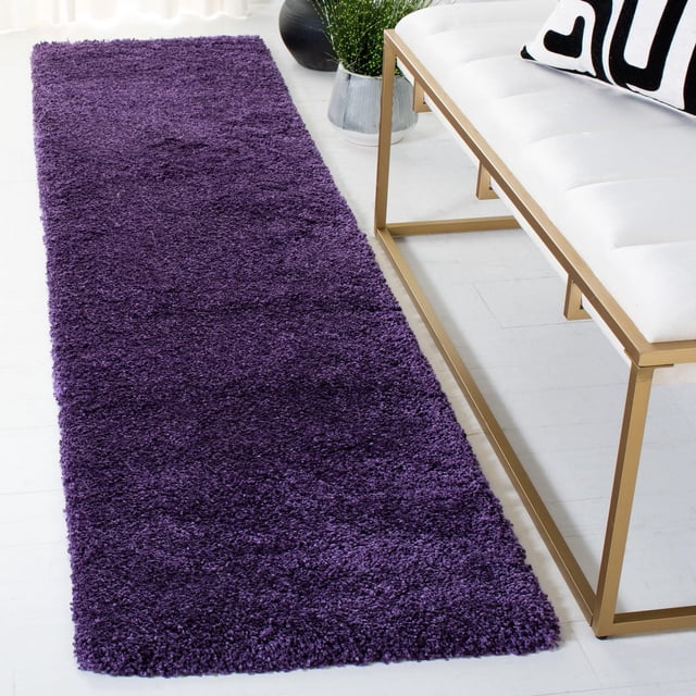 Safavieh Milan Harlow Solid Shag Area Rug in rich purple with dense synthetic yarns, placed on a light hardwood floor next to a white tufted bench with gold metal frame and a black and white geometric cushion, ideal for adding texture and warmth to contemporary home décor.