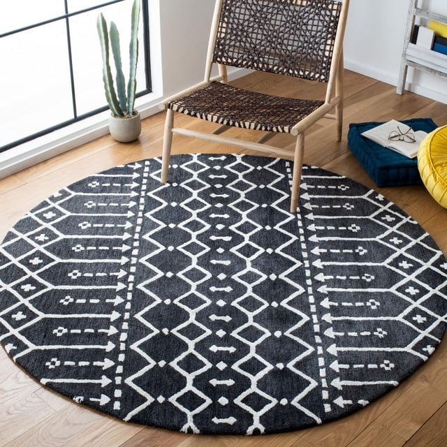 Safavieh Himalaya Darin Geometric Area Rug, a round accent rug with a black and white geometric pattern made from 100 percent hand-loomed wool, placed on a light wood floor under a woven wooden chair near a window with a potted cactus and a blue floor cushion with an open book and glasses.
