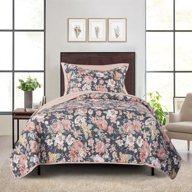 Mainstays Quilt-in-a-Bag Bedding Set on a twin bed with a dark wood headboard in a bright room. The lightweight quilt and matching pillow sham feature a floral pattern with pink, peach, cream, and green flowers on a dark background, bordered by solid light pink edges. Made from a soft recycled polyester blend, this easy-care set includes a quilt, pillow sham, and flat sheet. The room has large windows, a beige armchair, a side table with green foliage, and three framed botanical prints above the bed.